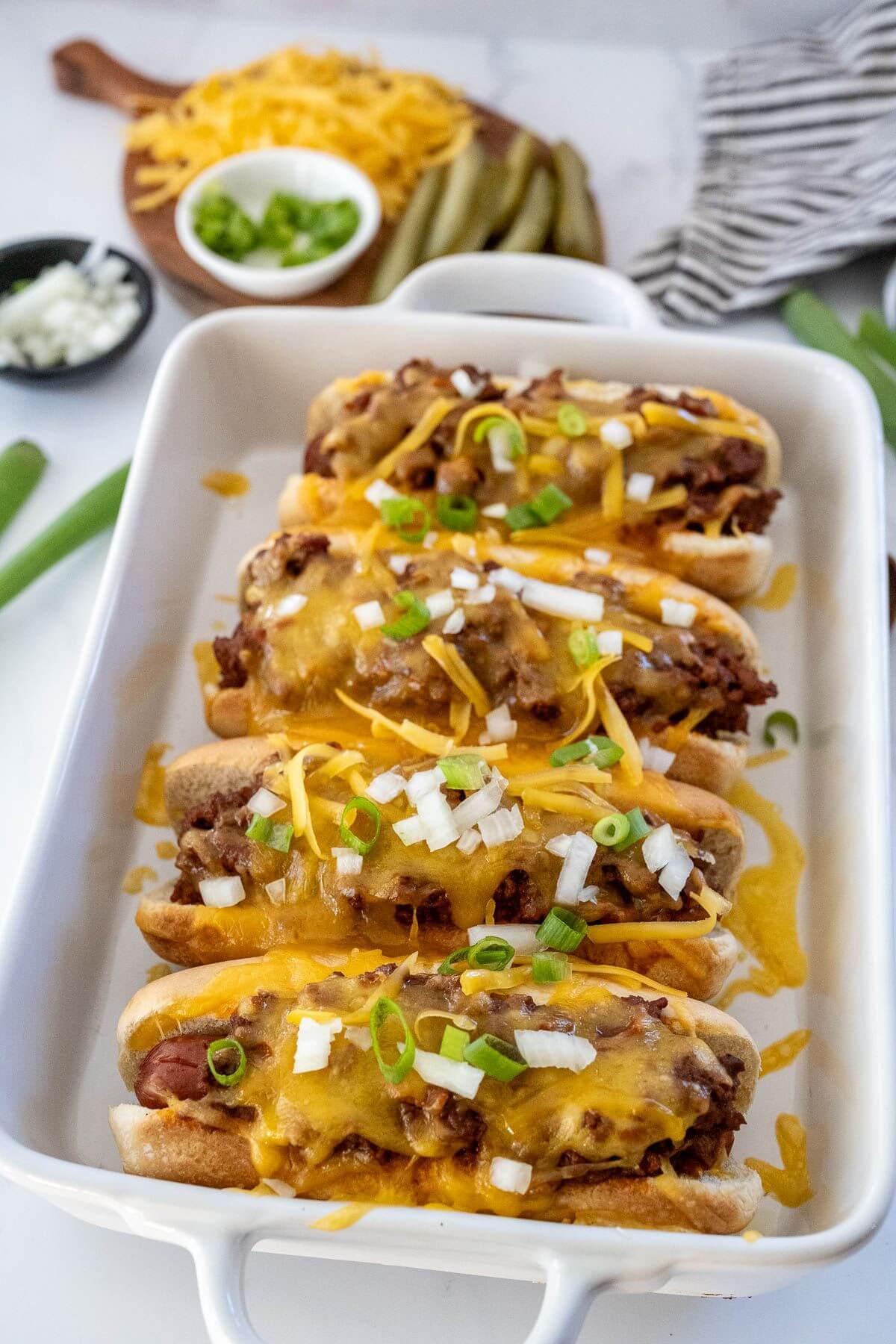 A baking dish full of chili dogs with cheese, onions, and green onions is next to other toppings.