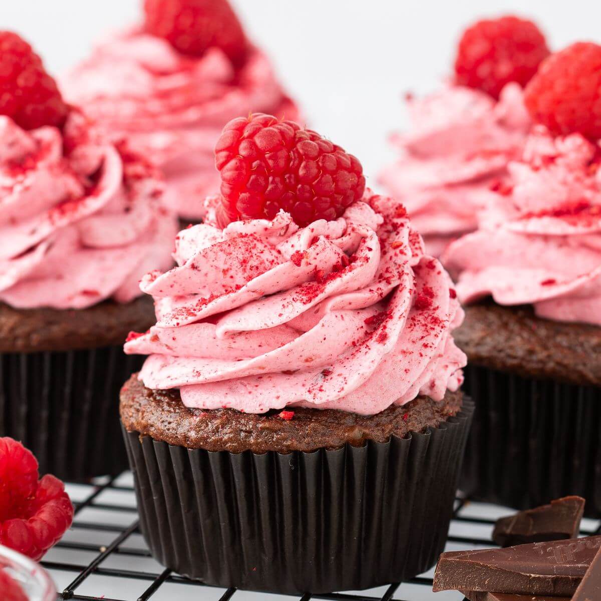 Five cupcakes topped with pink thick icing, red sprinkles, and raspberries sit on a wire rack.