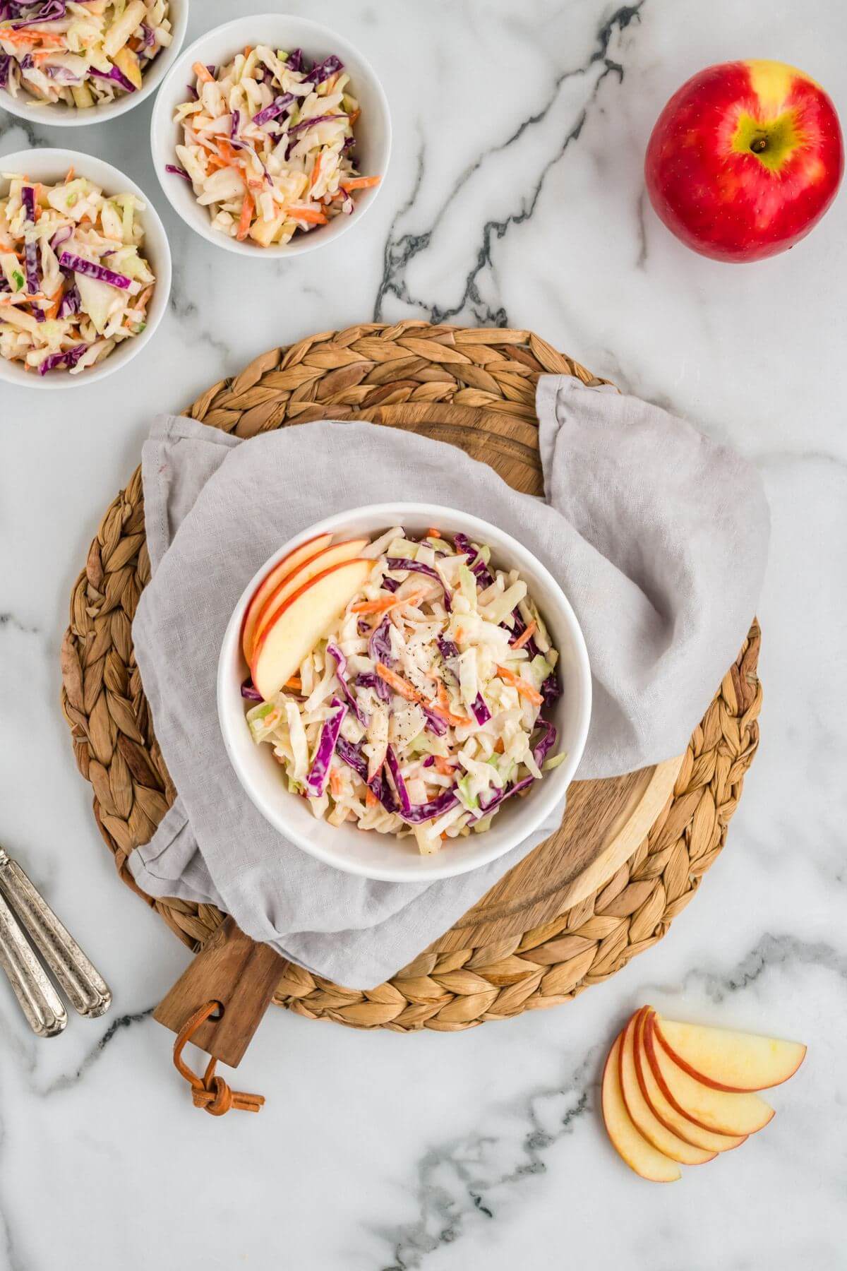 One bowl of coleslaw is centered on a wicker platter with more bowls and apples behind it.