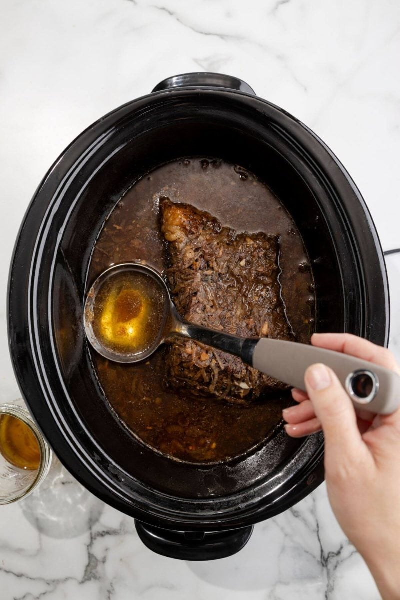 A hand dips a ladle into broth in a crockpot holding the brisket.