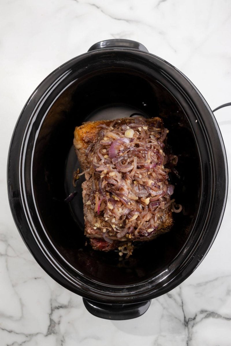 A mix of onions and other seasonings sit on top of brisket in an empty crockpot.