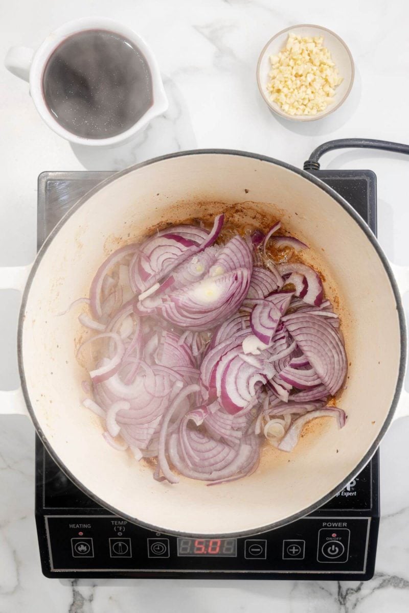 Onions saute in a pan on an electric stovetop with other ingredients behind it.
