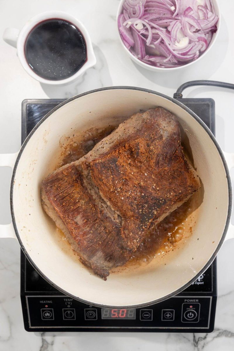 A brisket is cooking and browning in a pan on an electric stovetop.