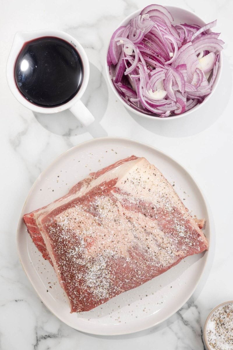 A seasoned raw brisket in on a plate next to sauces and raw onion slices.