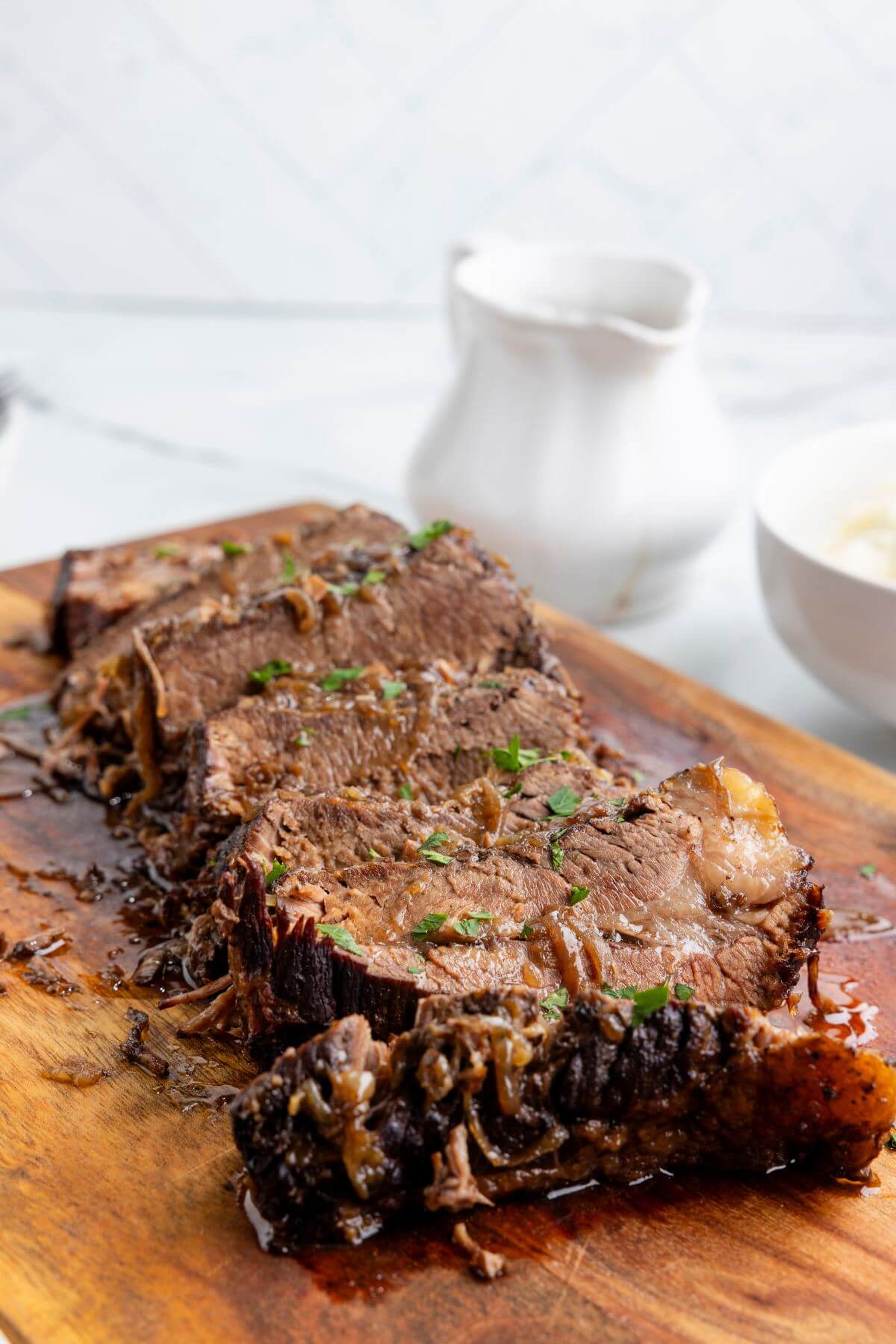 A gravy boat sits behind a wooden board serving brisket.