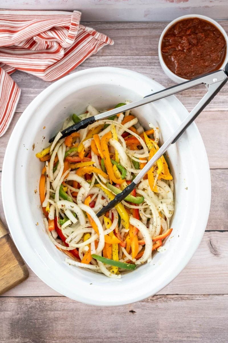 Tongs rest on top of a crock pot filled with colorful vegetables next to bowl of marinara sauce.