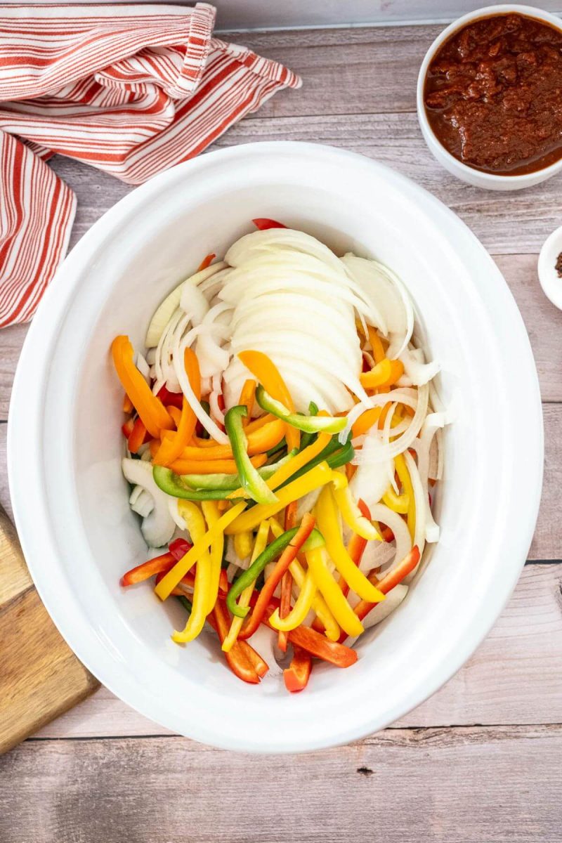 Bright yellow, green, and red bell pepper strips and onion slices fill a crock pot.