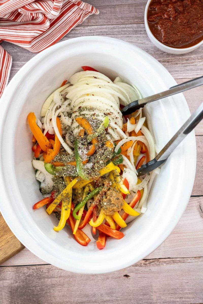 Tongs sit at the edge of a white crock pot filled with colorful uncooked bell pepper and onion slices.