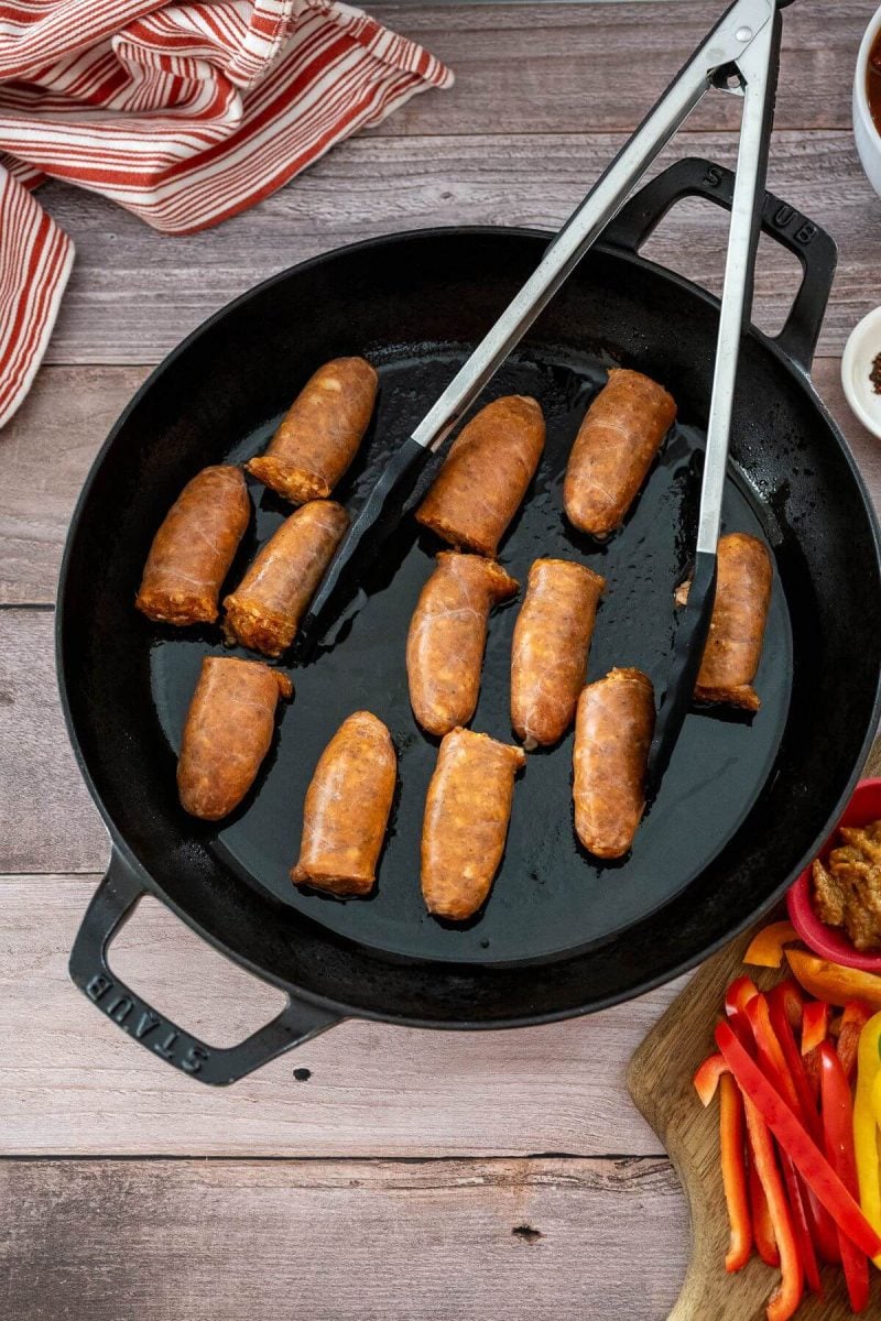 A pair of tongs goes to pick up a sausage from the cast iron pan.