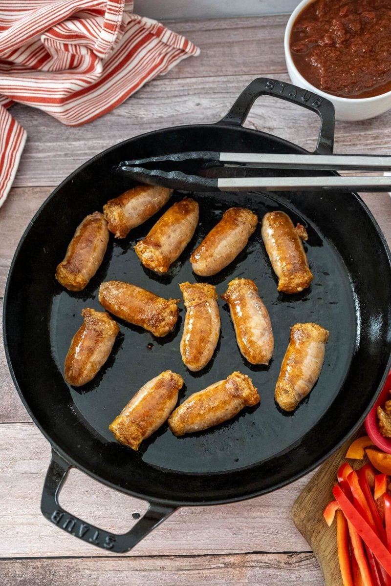 A cast iron pan has sausages browning in it and a pair of tongs at the ready.