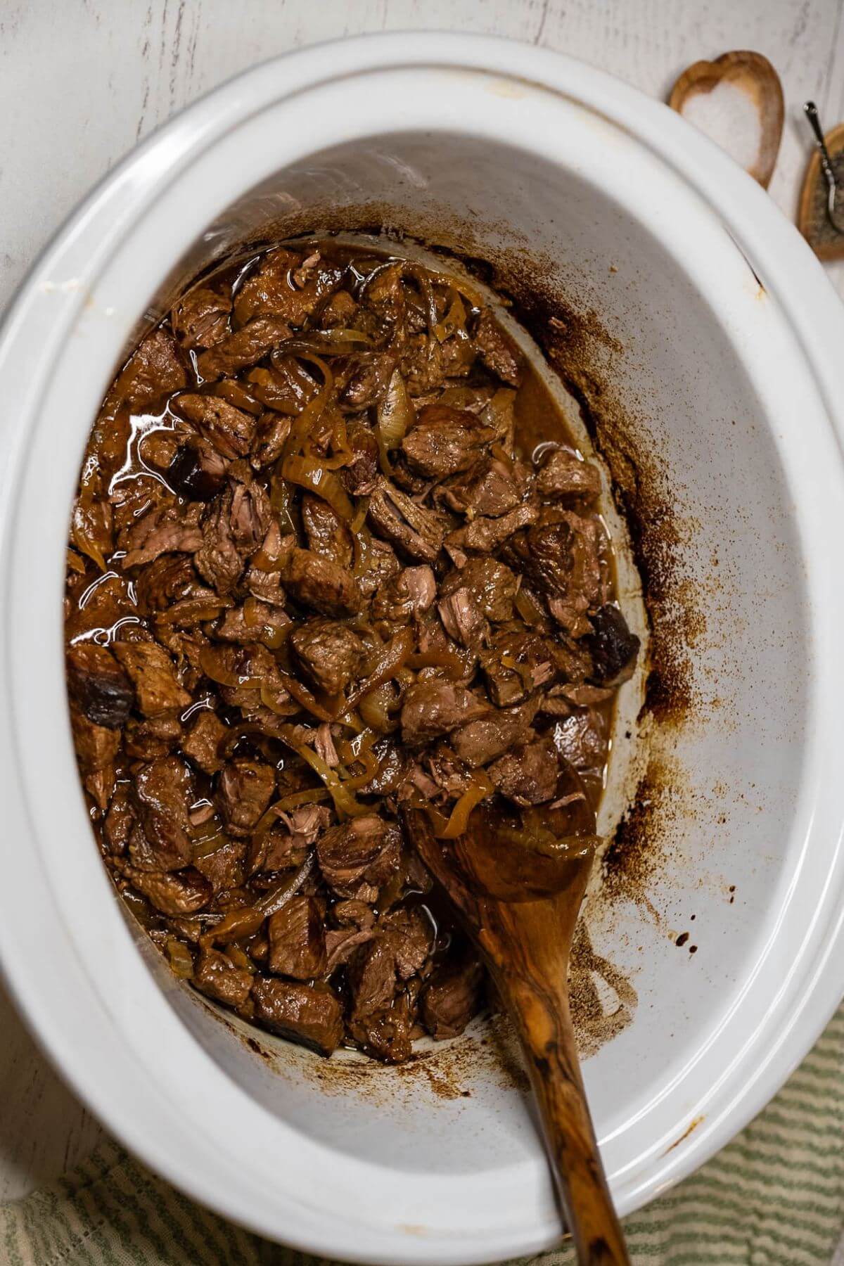 An open crock pot shows steak chunks, onions, and sauce stirred by spoon.