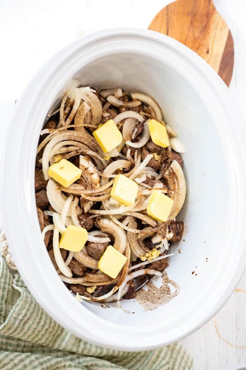 A lidless crock pot shows uncooked onion slices, meat, and butter chunks.