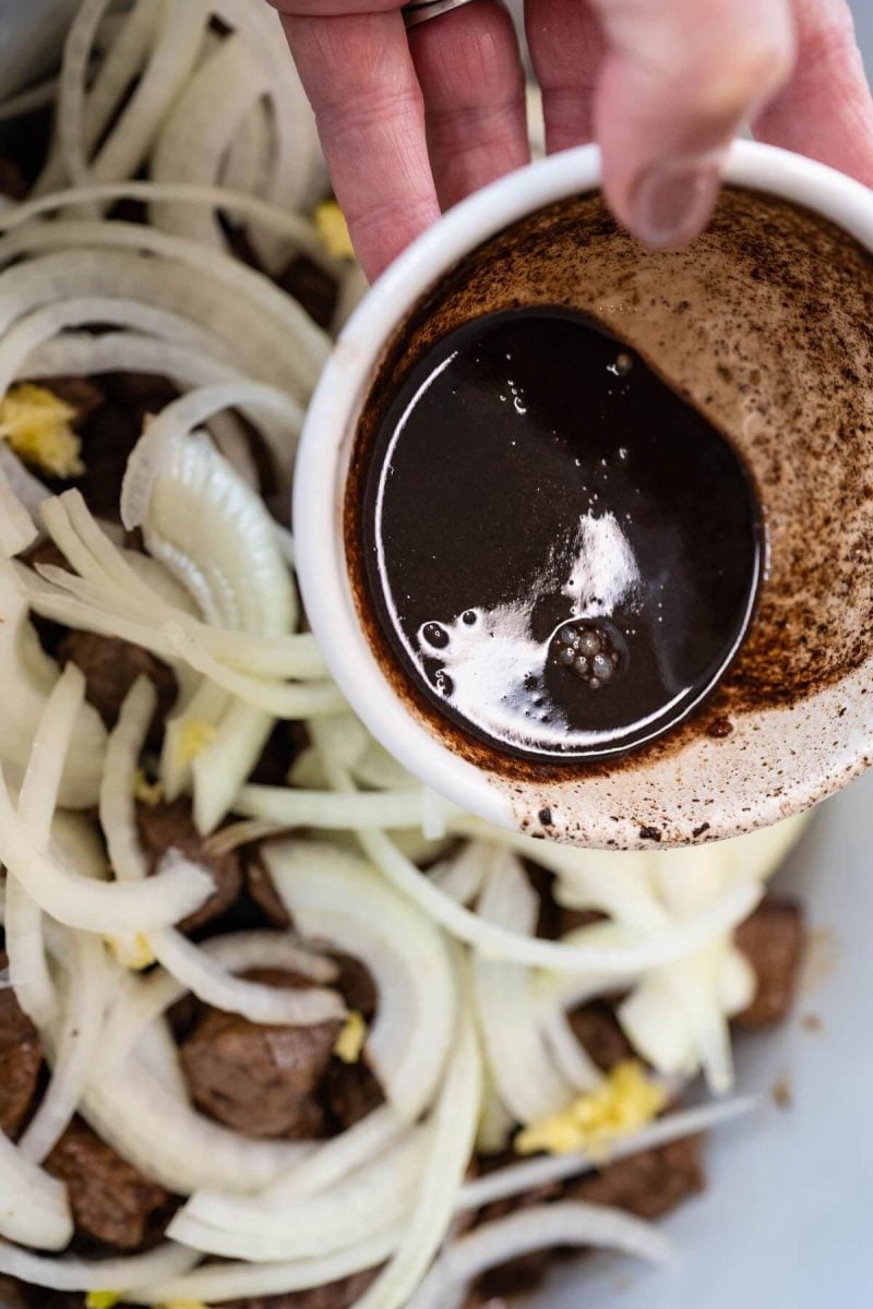A hand pours brown thick liquid out of small bowl into crockpot onions.