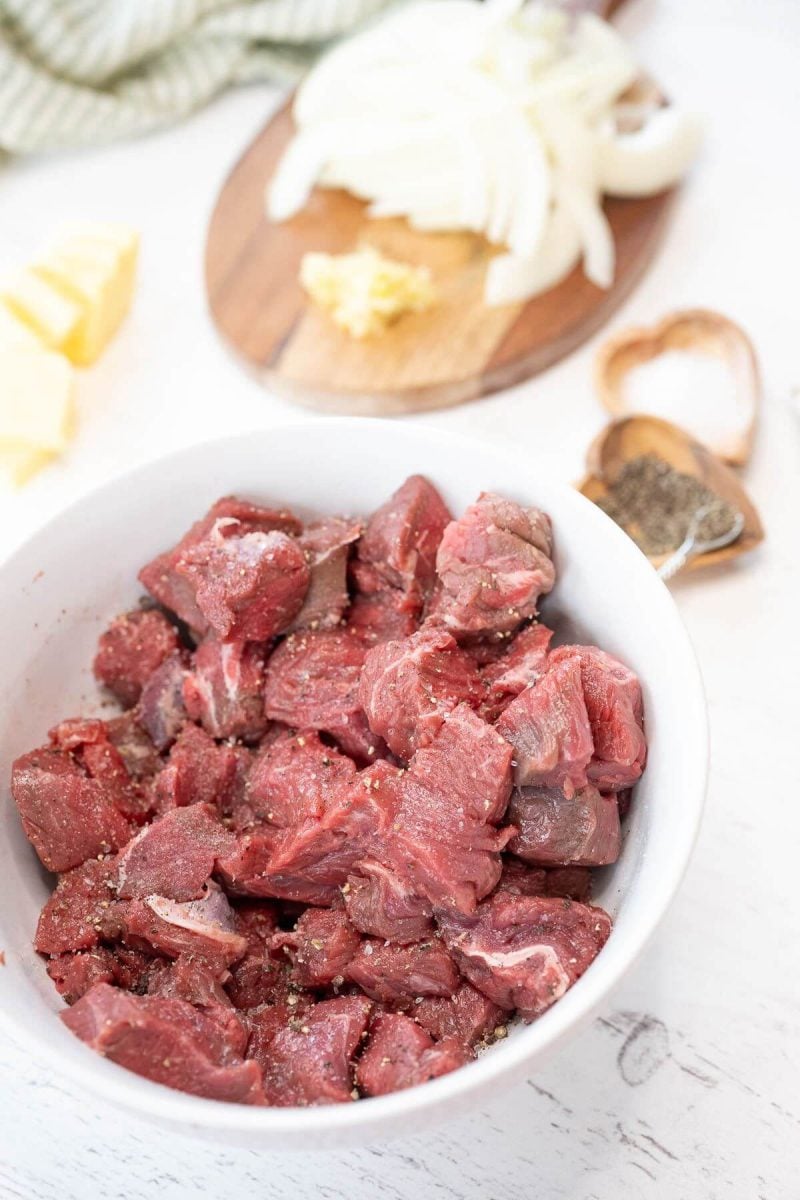 Raw chunks of meat in a bowl are sprinkled with seasonings.