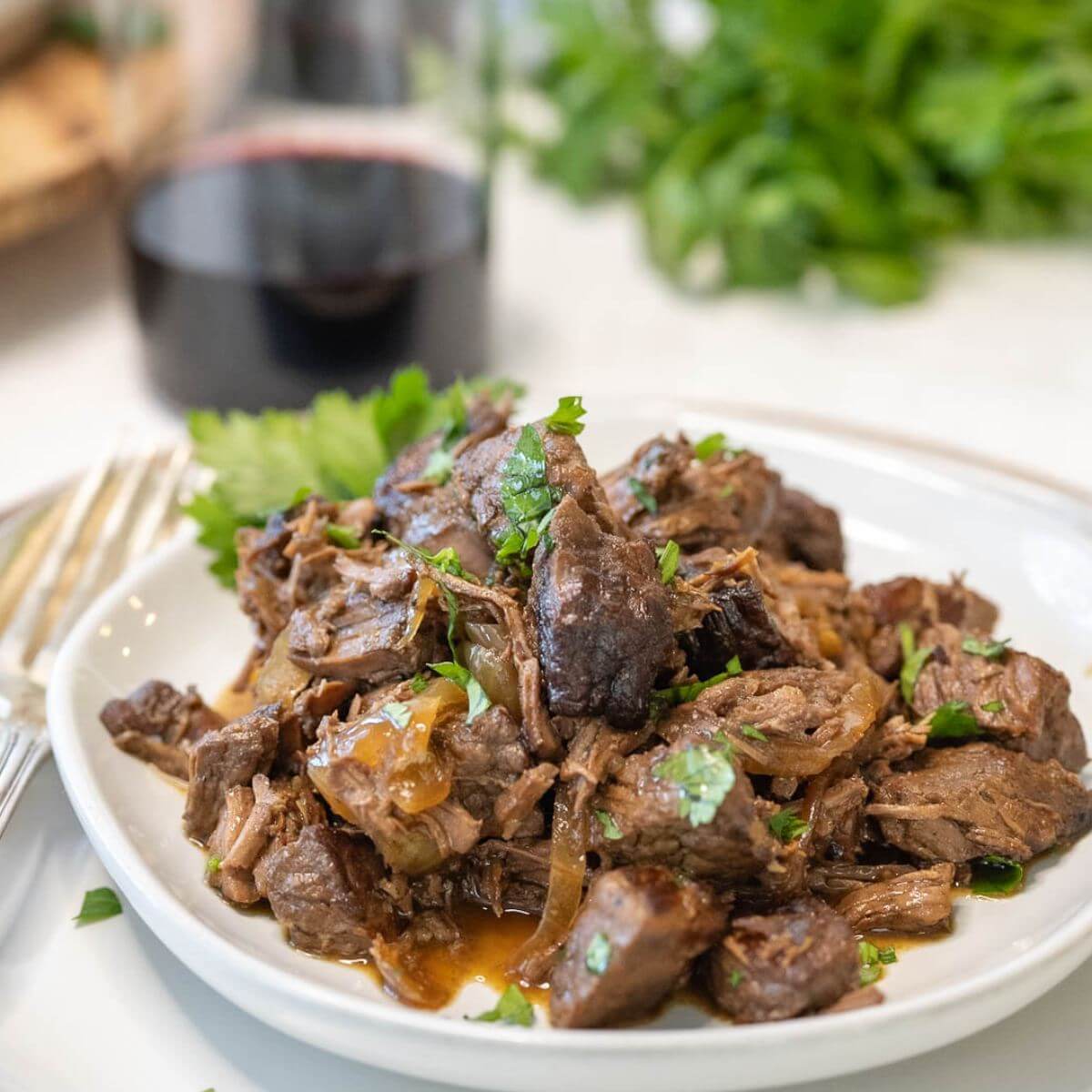 A plate is filled with steak bites and onions with wine glass and greens behind it.