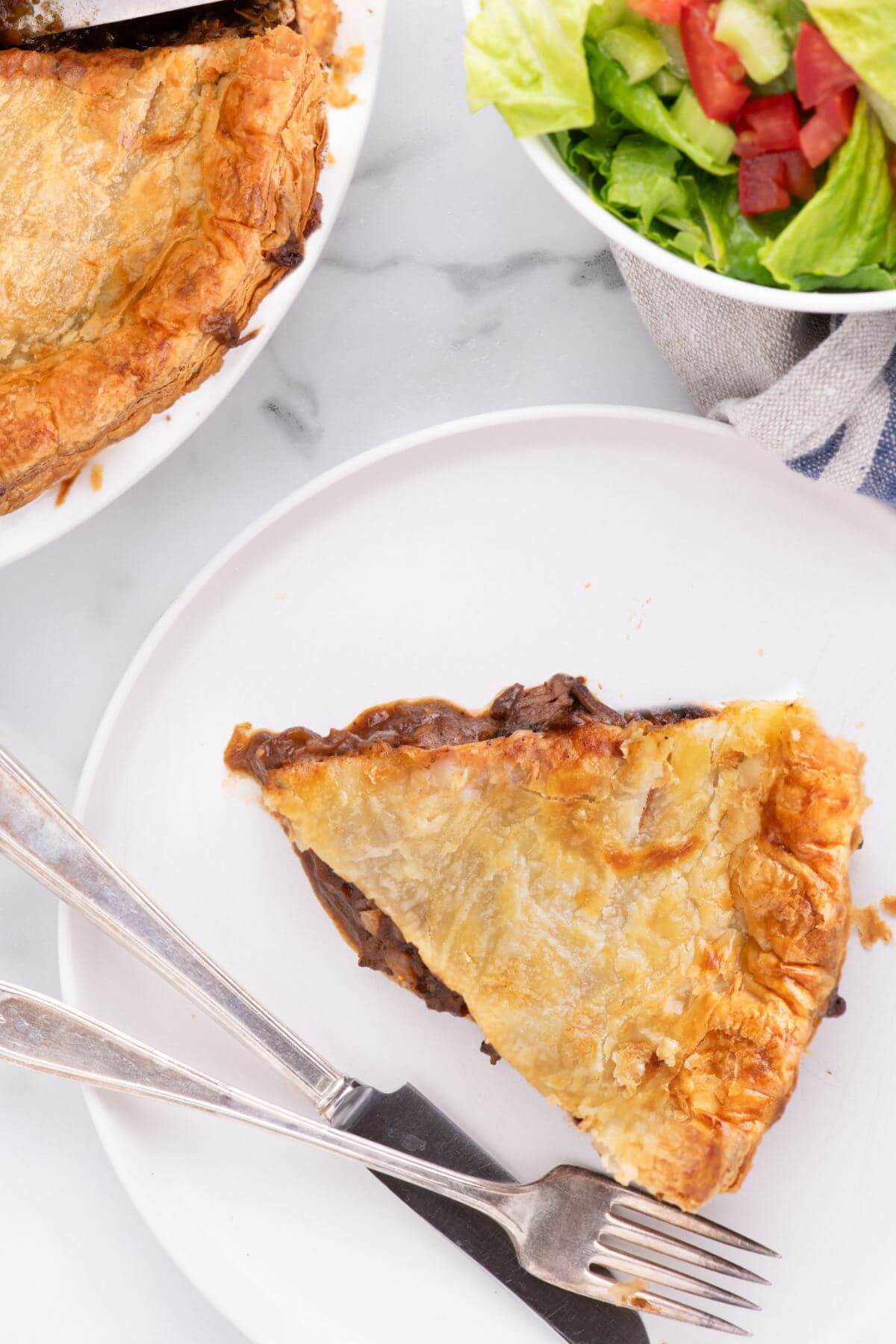 A fork and knife lay by a golden flaky steak pie slice and salad.