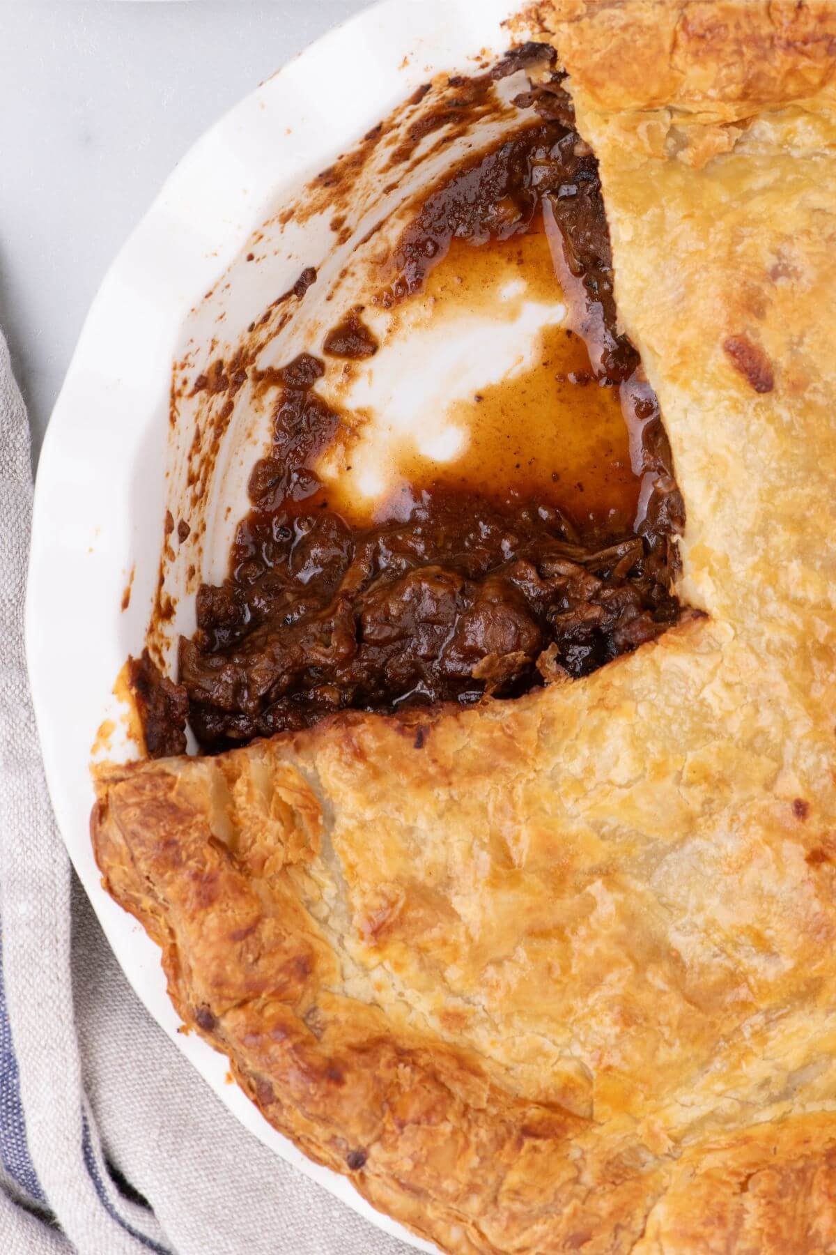 Irish steak and ale pie with beefy filling with gravy spilling out from under the puff pastry crust in a pie plate. 
