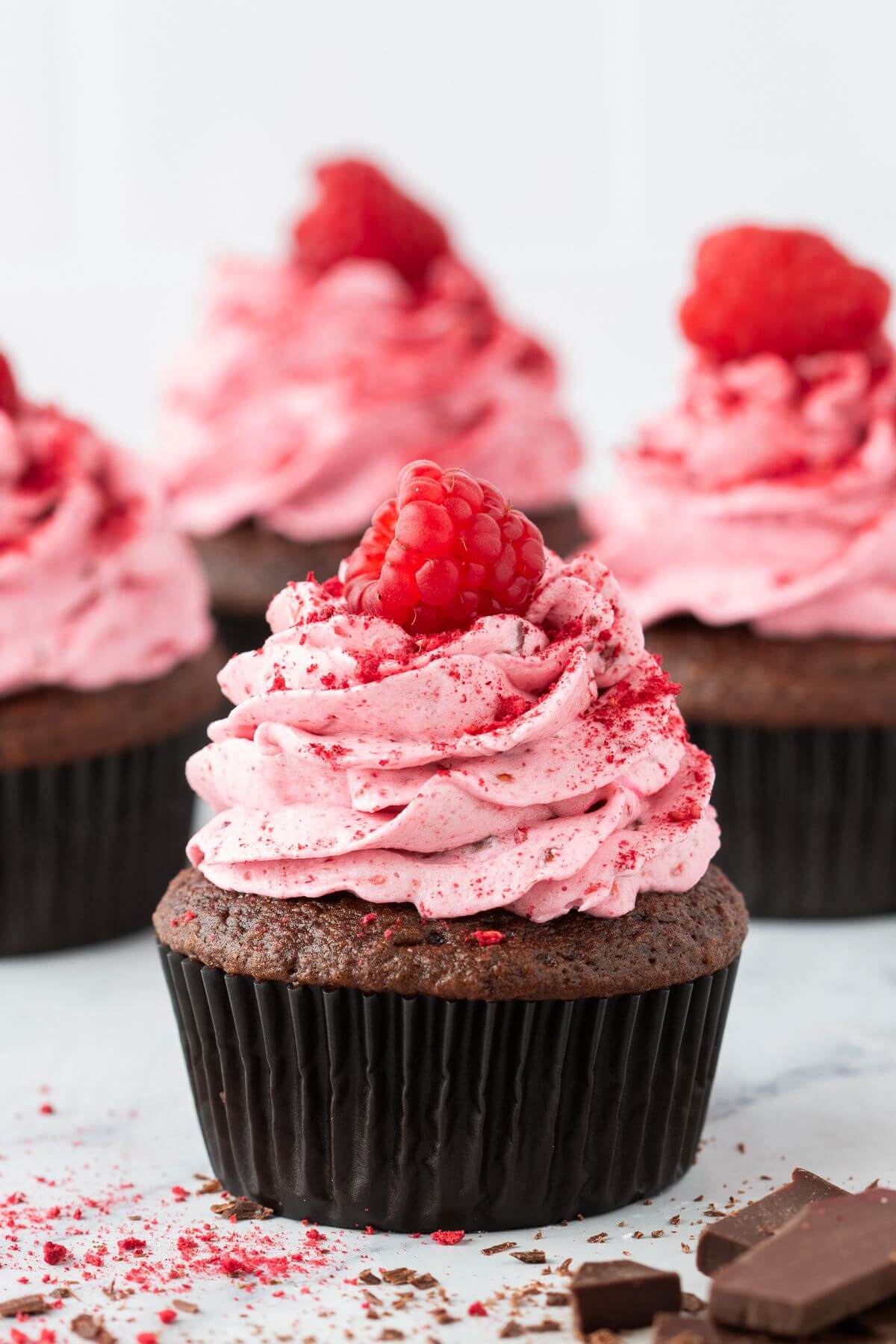 Four pink frosted cupcakes sit on table scattered with red sprinkles and chocolate chunks.