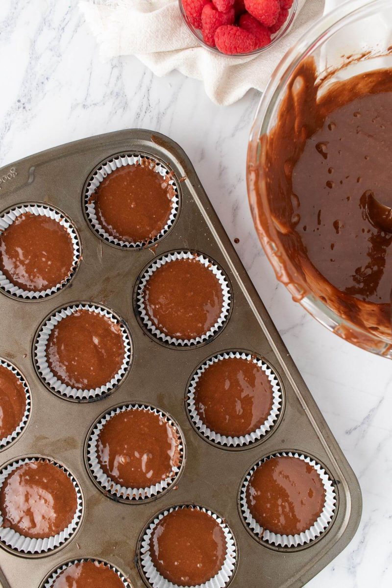 A cupcake baking pan is filled with chocolate batter next to the bowl of batter.