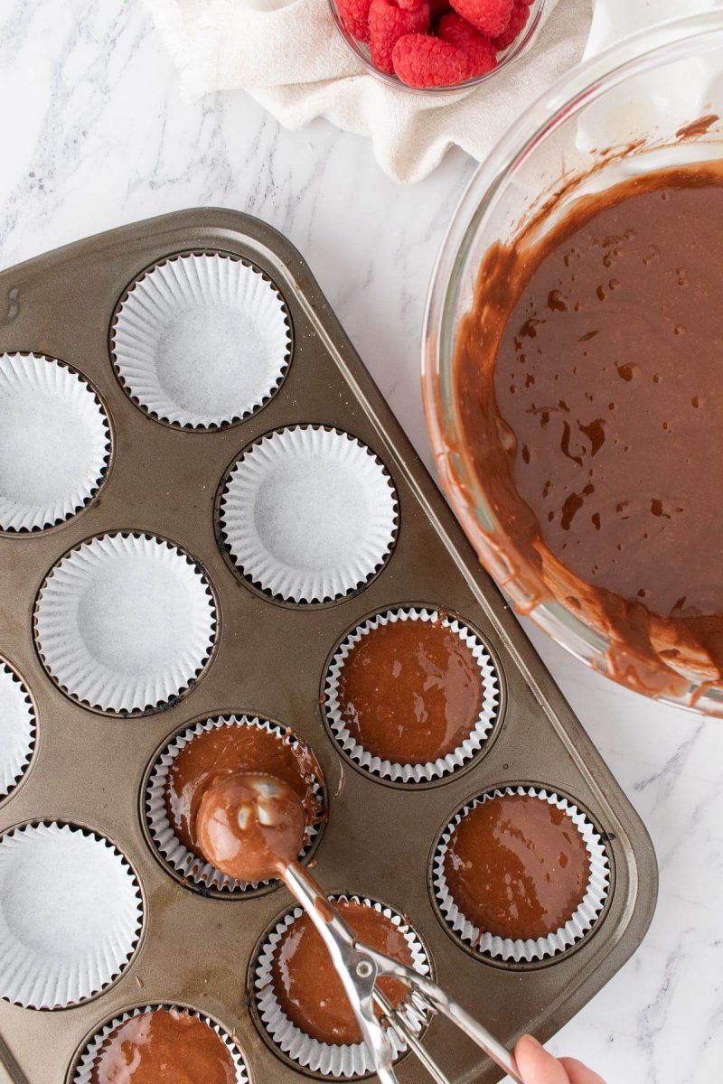 A hand uses a metal scoop to deposit chocolate batter into cupcake liners in pan.