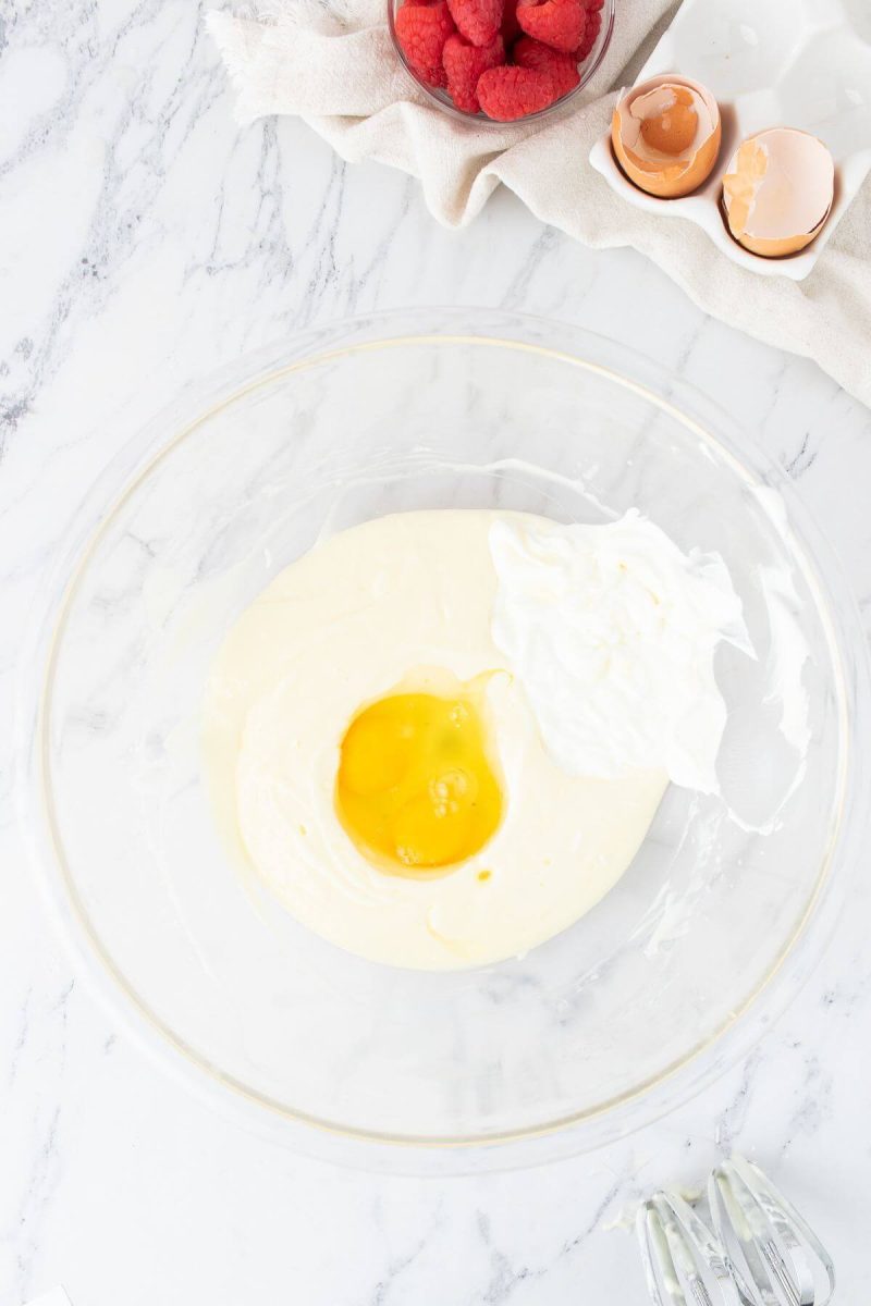 Two eggs and white sour cream sit on top of other wet ingredients in bowl.