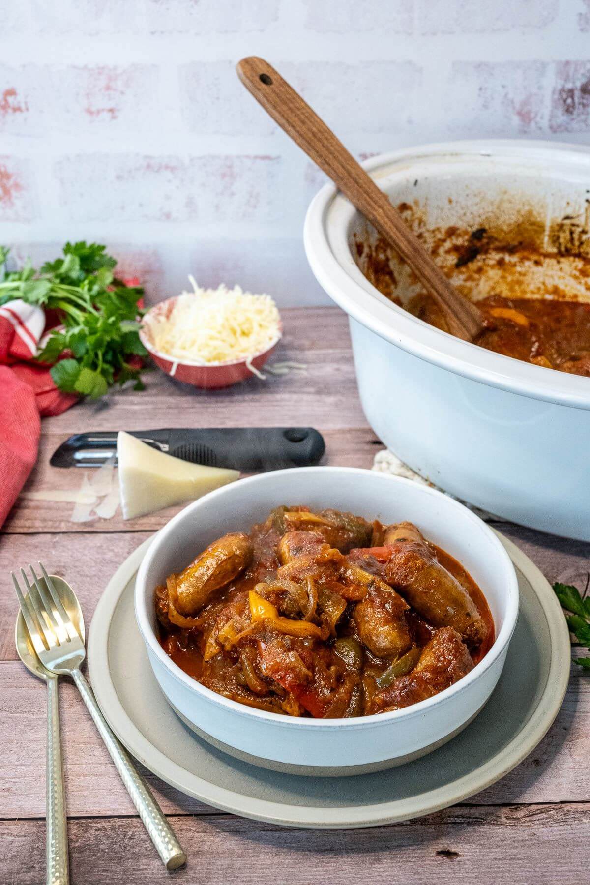 A fork lies on a spoon next to a filled bowl of sausage and pepper mix by its crock pot.
