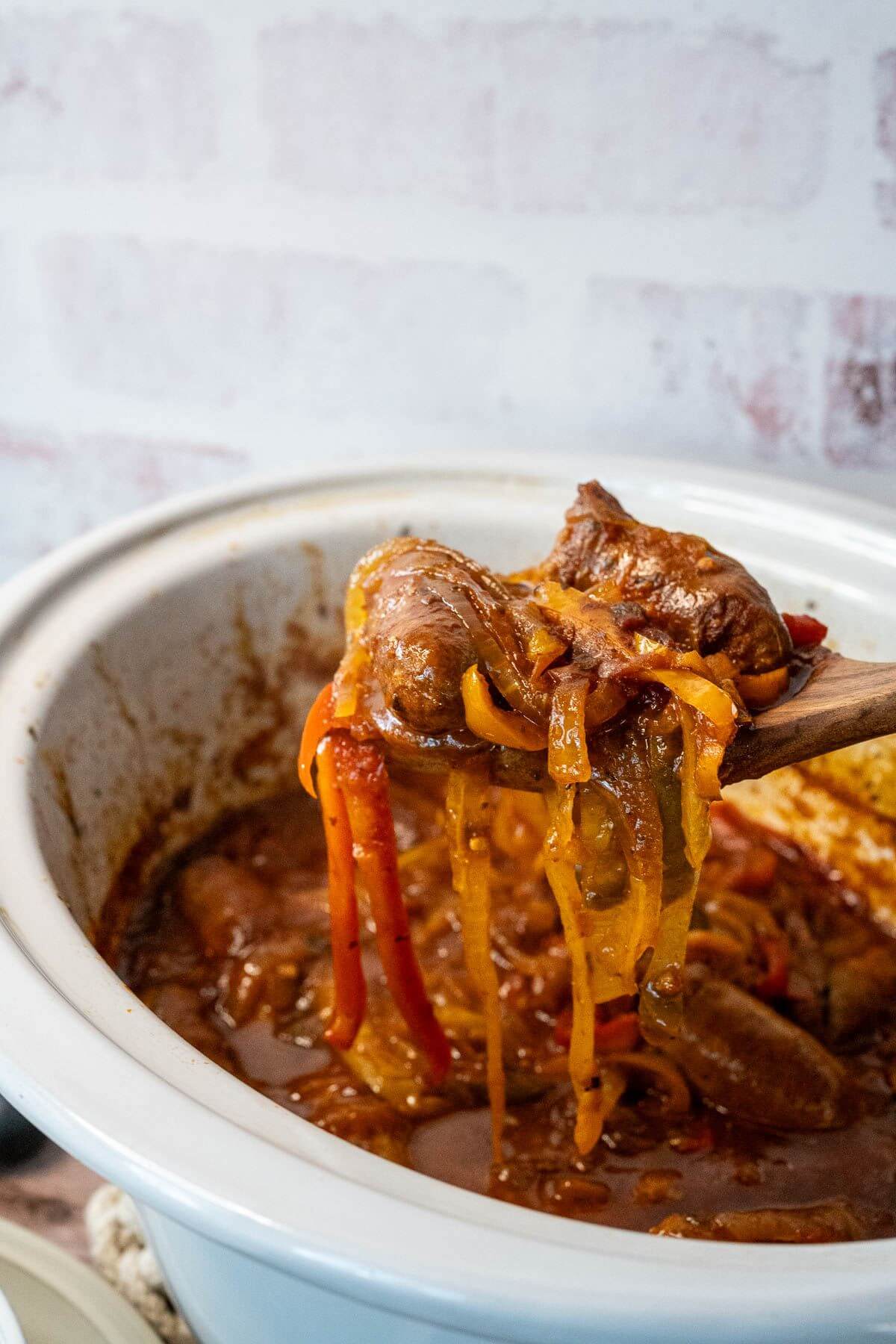 A wooden spoon lifts up a huge collection of pepper strips and a couple of sausages out of crock pot.