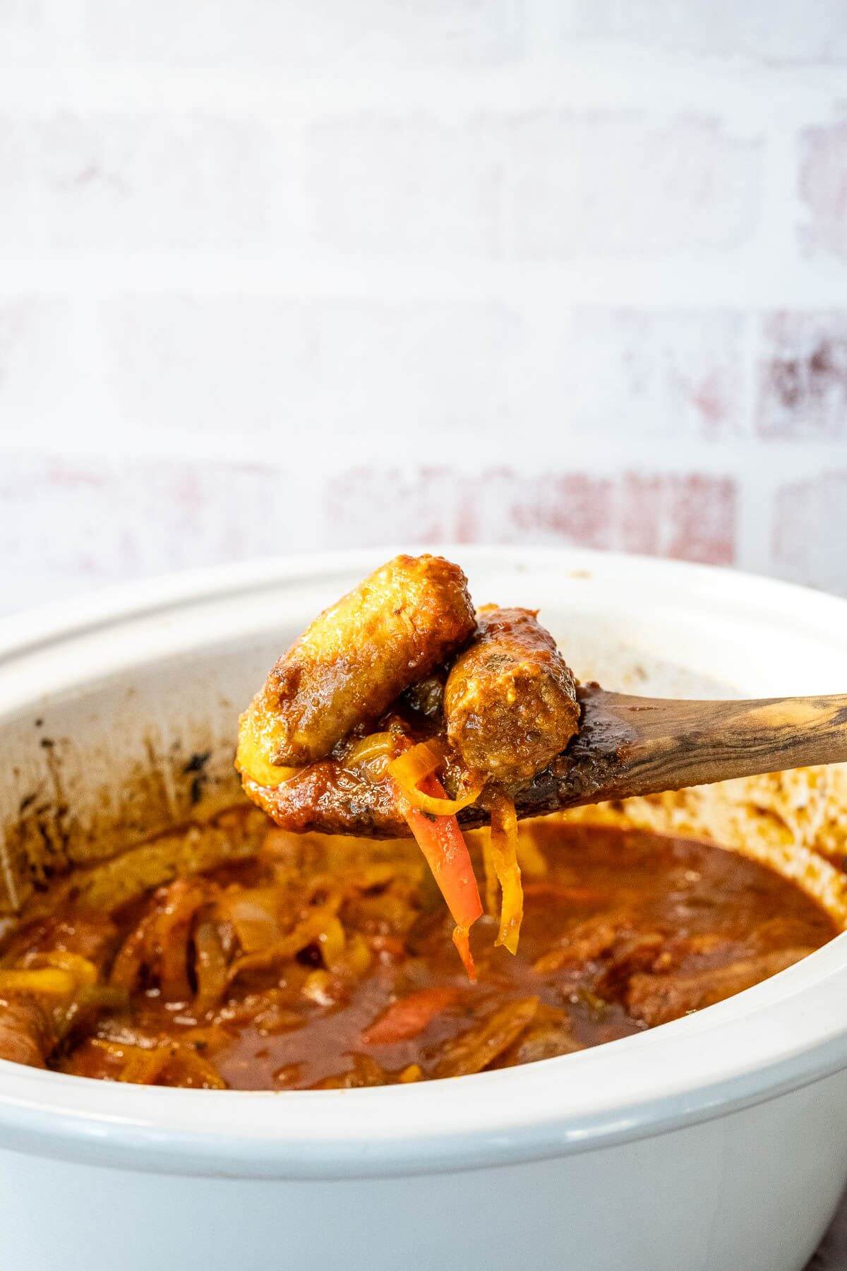 A wooden spoon lifts up two sausages and some peppers from crock pot.