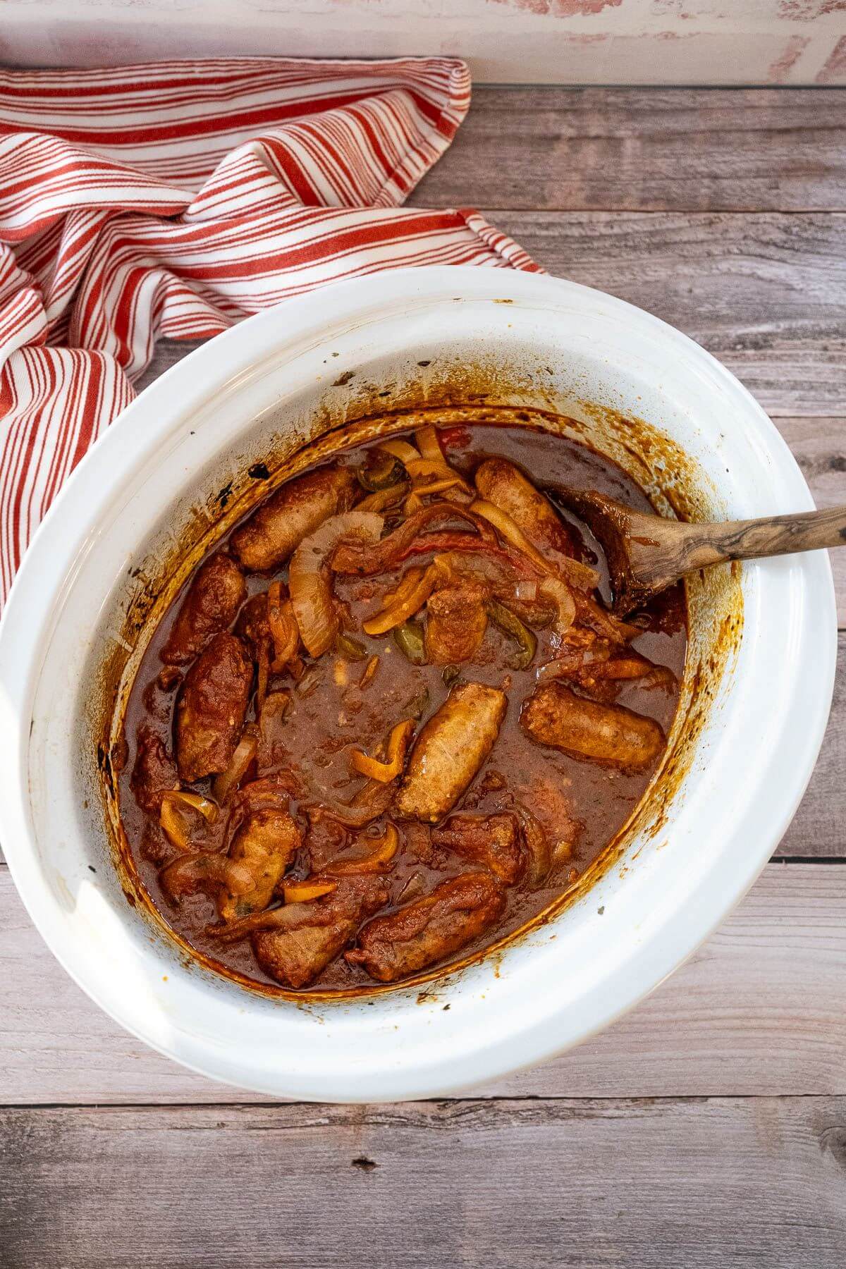 A white crock pot is shown from above so the sausages and peppers inside are visible.