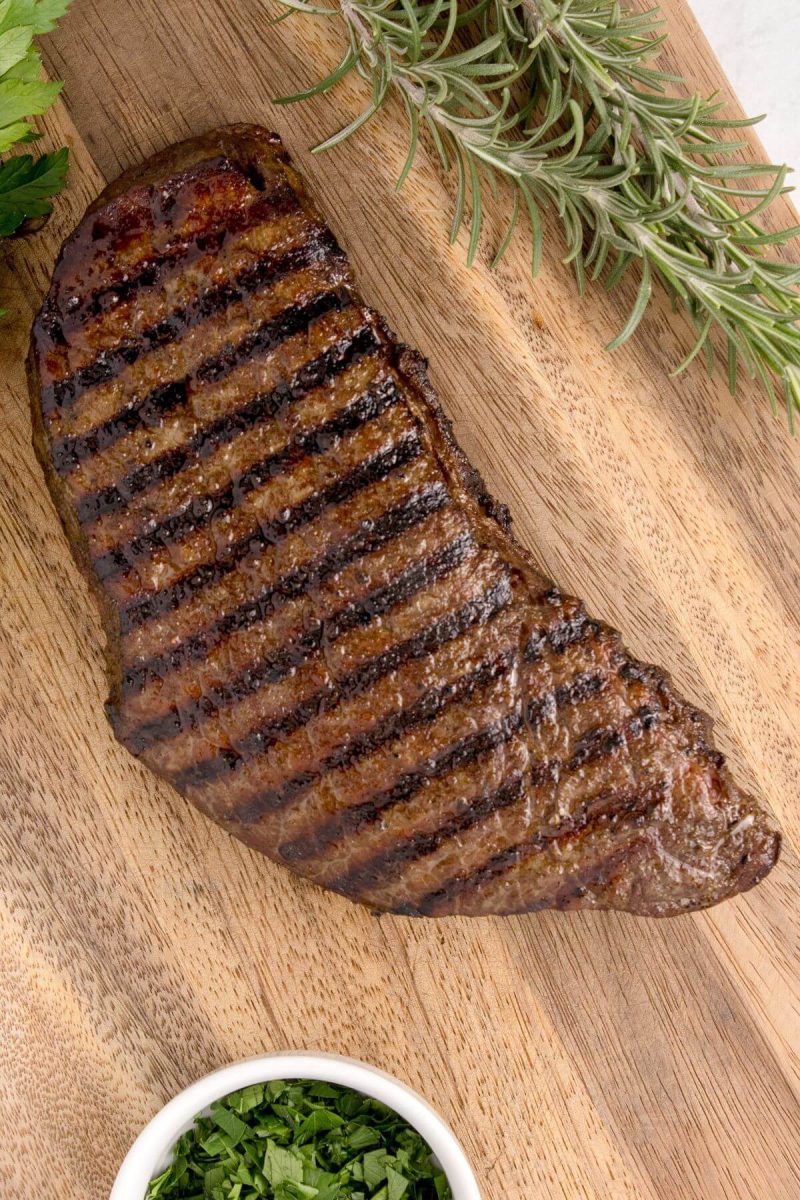 Steak with grill marks is on a wooden board by rosemary and greens.