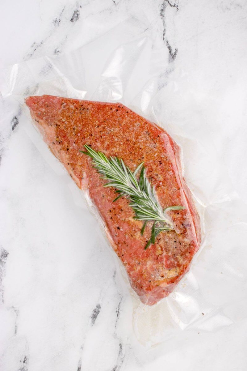Sprig of rosemary is resting on a meat hunk in a plastic bag.