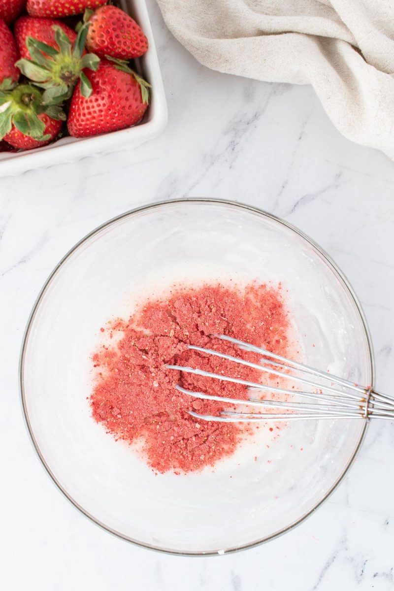 A whisk stirs pink powdery ingredients in a glass bowl.