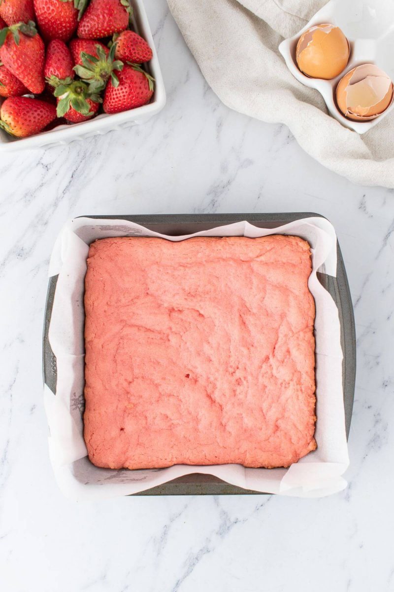 A pink baked cake is in a pan next to bowl of strawberries.