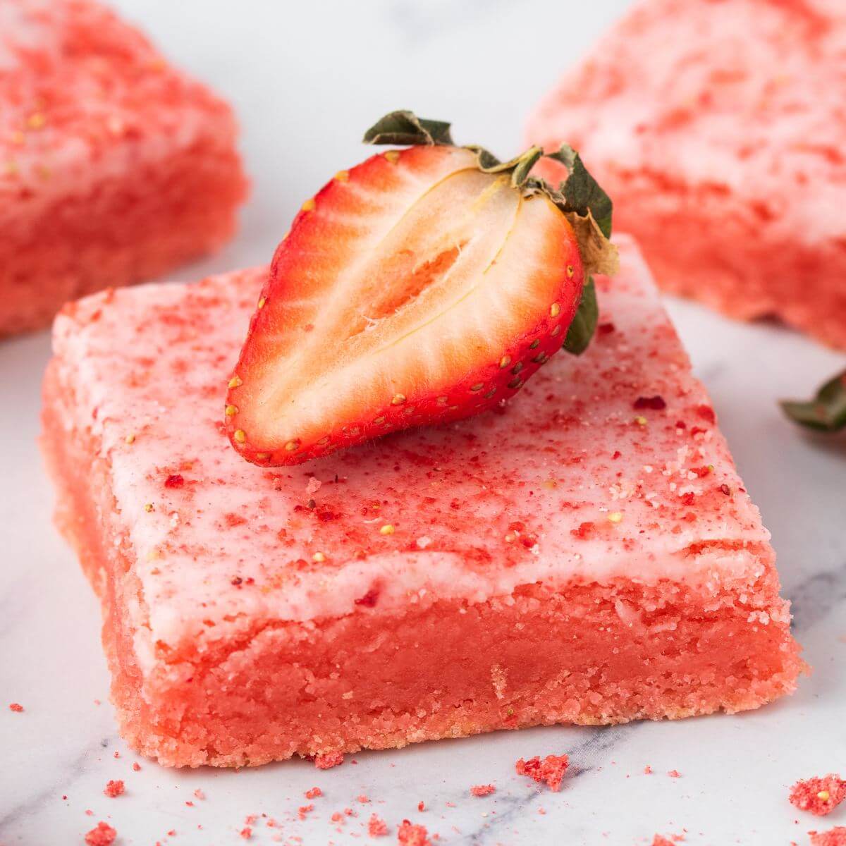 A strawberry rests on a pink baked square next to some crumbles.
