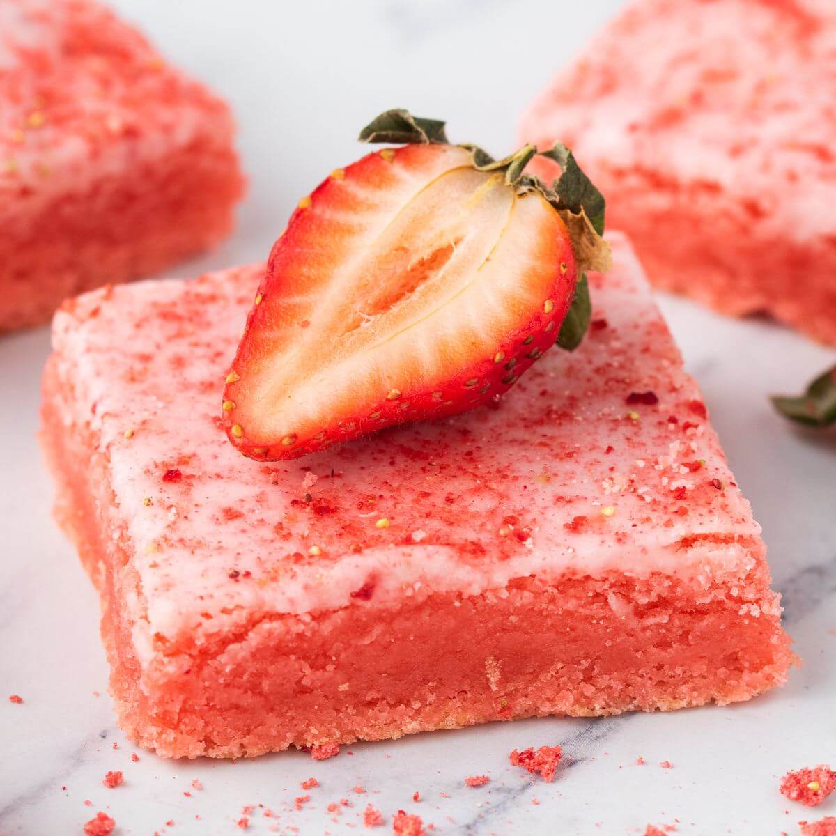 A strawberry rests on top of a frosted strawberry brownie next to some crumbles sprinkled around. 