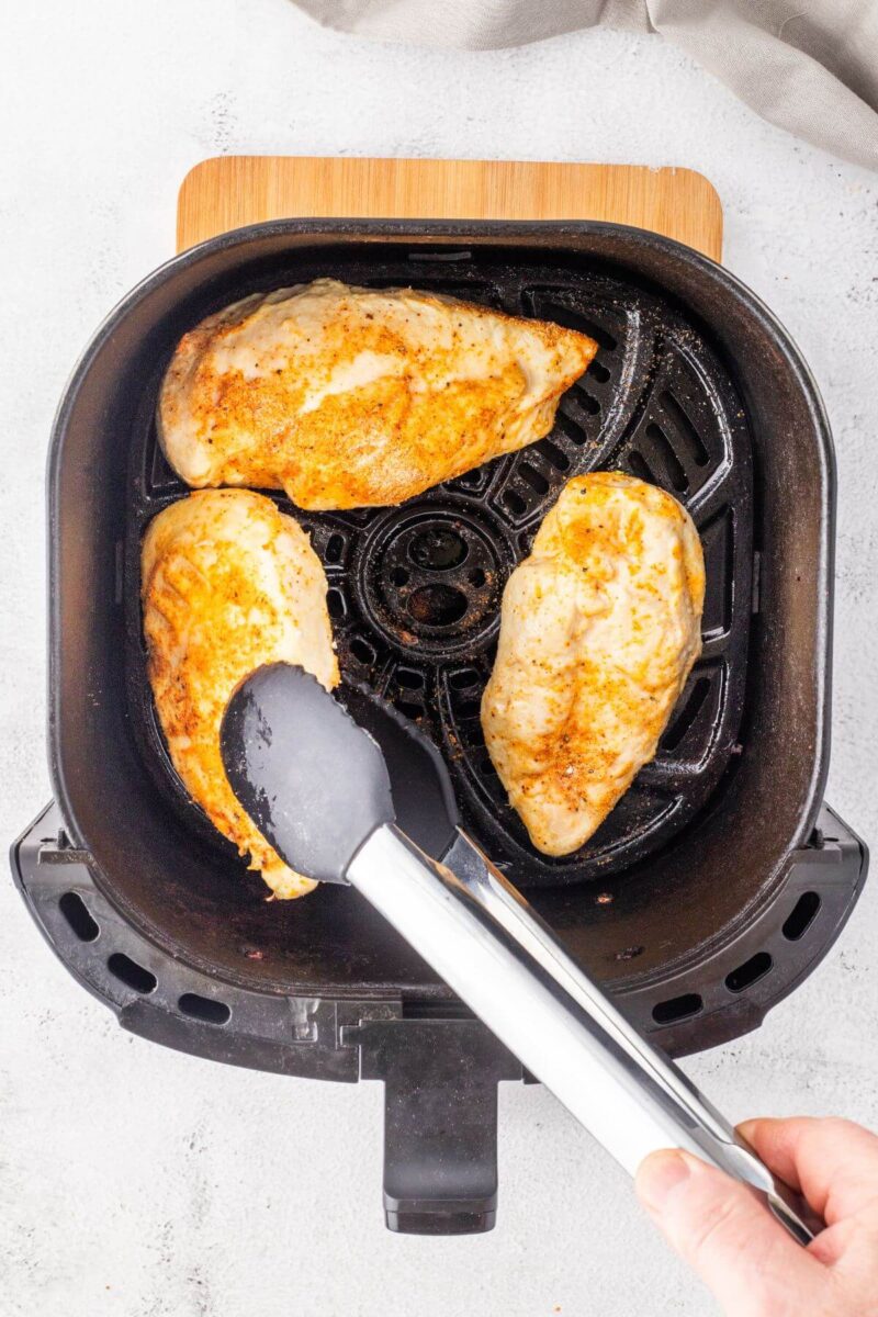 Tongs lift up a chicken breast to flip in air fryer device.