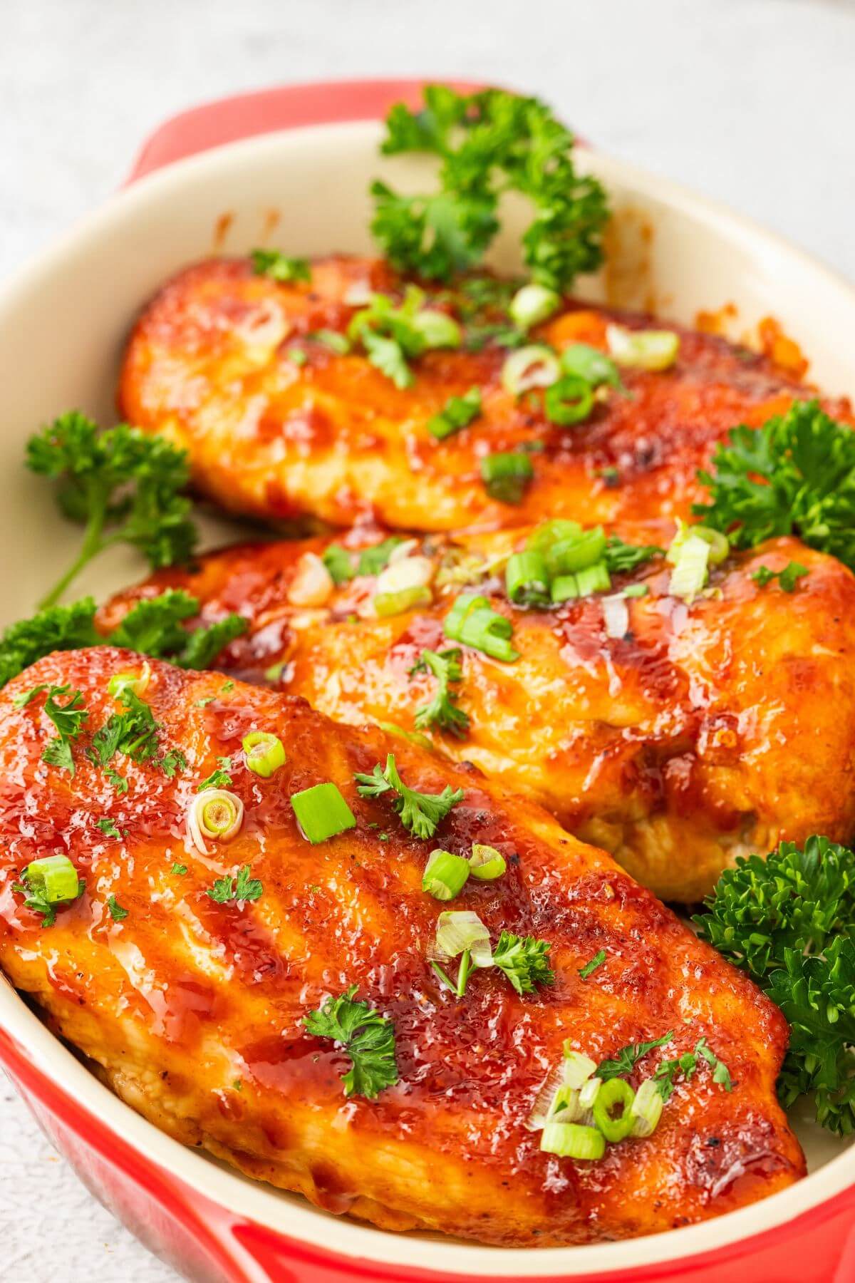 A red baking dish is filled with three cooked chicken breasts and covered in green onions and herbs.