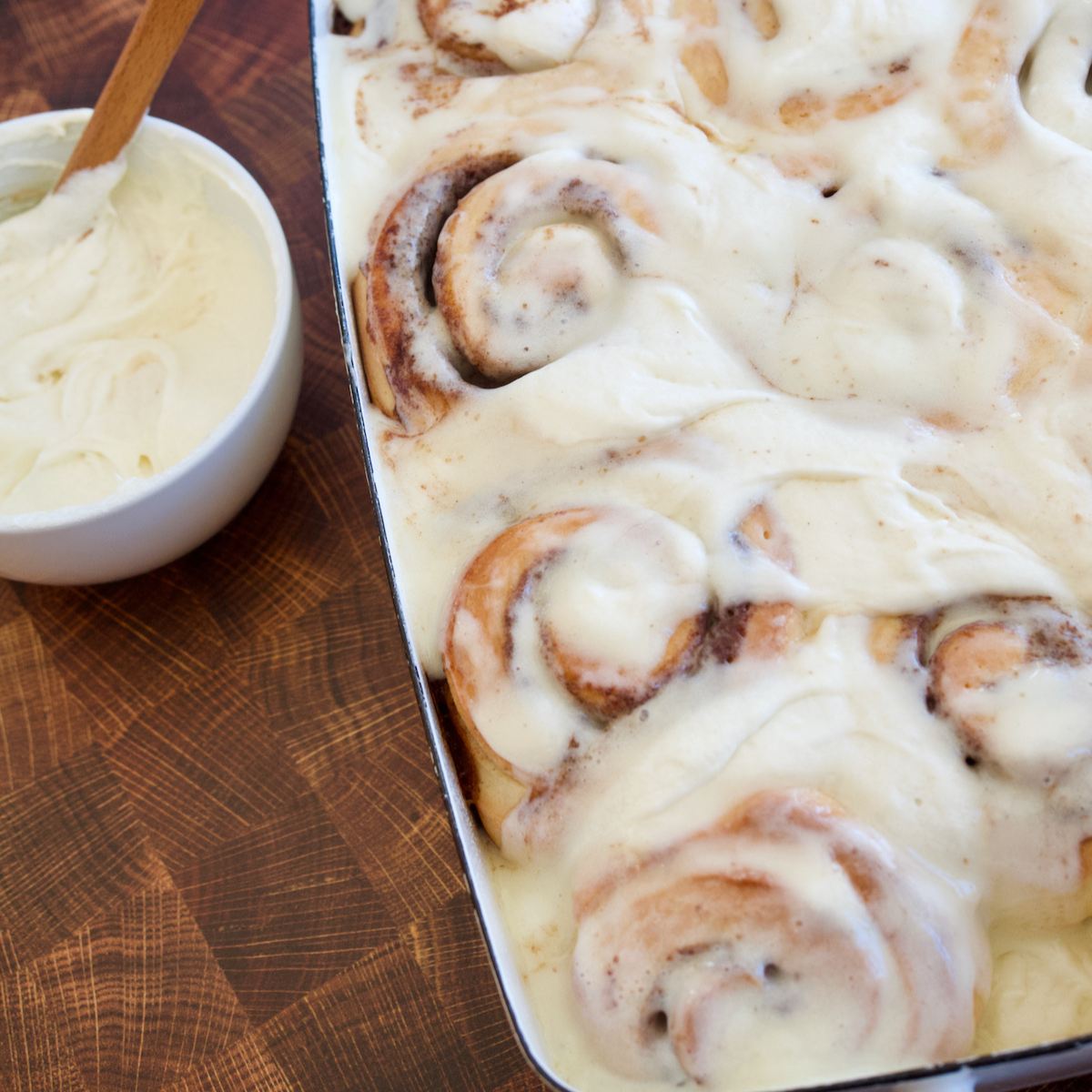 Pan of frosted gooey cinnamon rolls with bowl of frosting nearby.