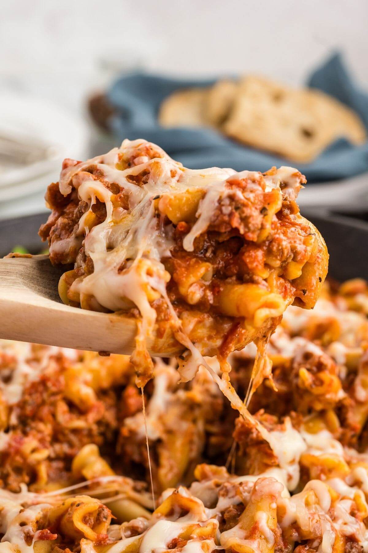 A wooden spoon holds up a pile of meaty, gooey cheese covered noodles above the pan.