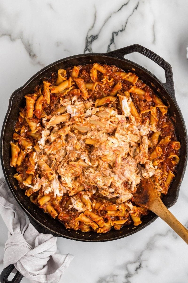 Ricotta cheese is pressed into the beef, noodle and tomato mix with a wooden spoon.