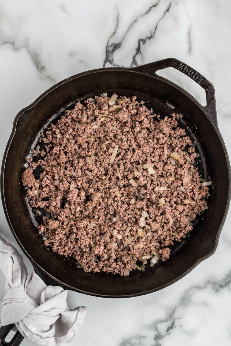 Ground beef cooks in an iron pan with minced onion.