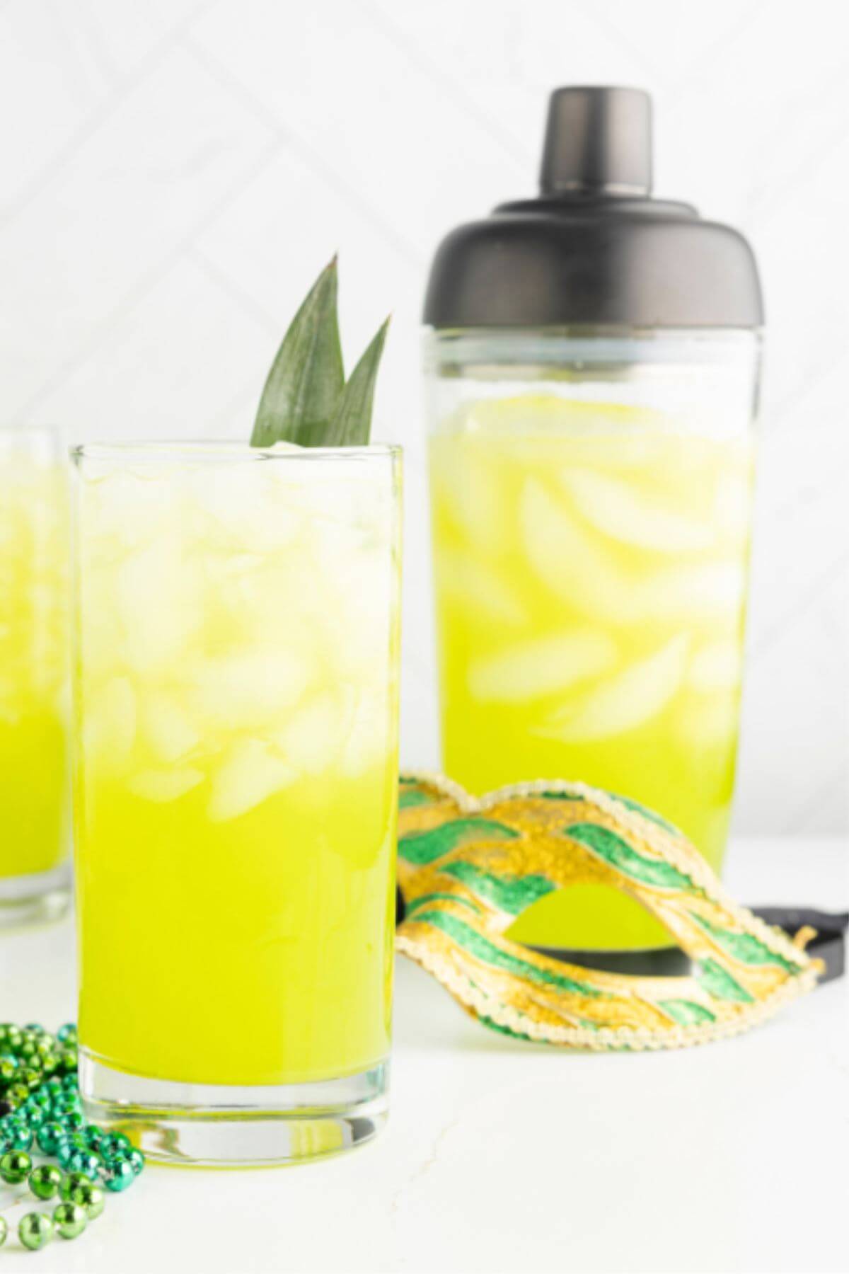 A gold and green mask and beads separate a yellow cocktail in glass from the cocktail shaker.