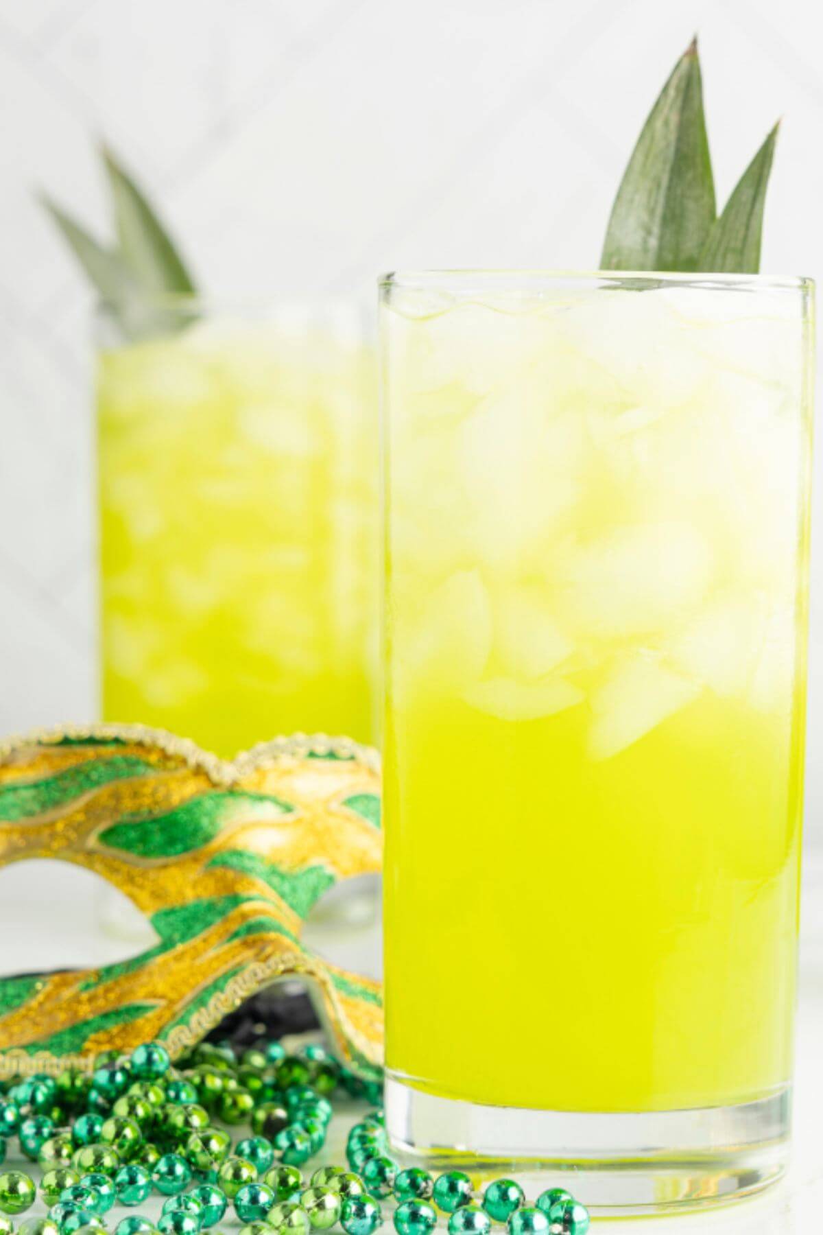 Two yellow cocktails with leaf garnish are next to green and gold mask and beads.