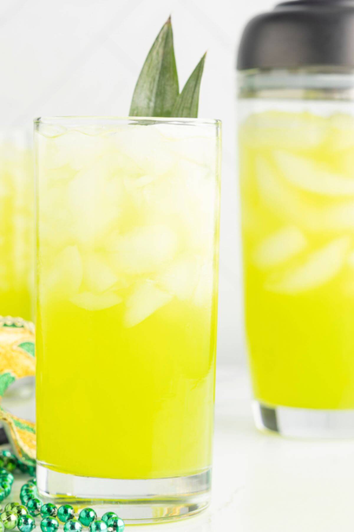 A shaker cup of yellow cocktail mix is next to glasses full of beverage.