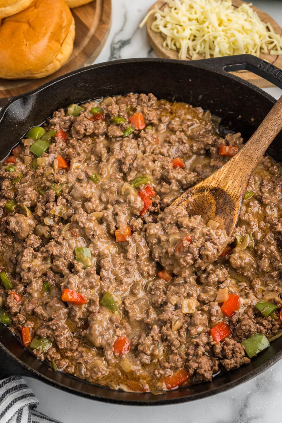 A wooden spoon dips into meat, veggie, and sauce mixture in skillet.