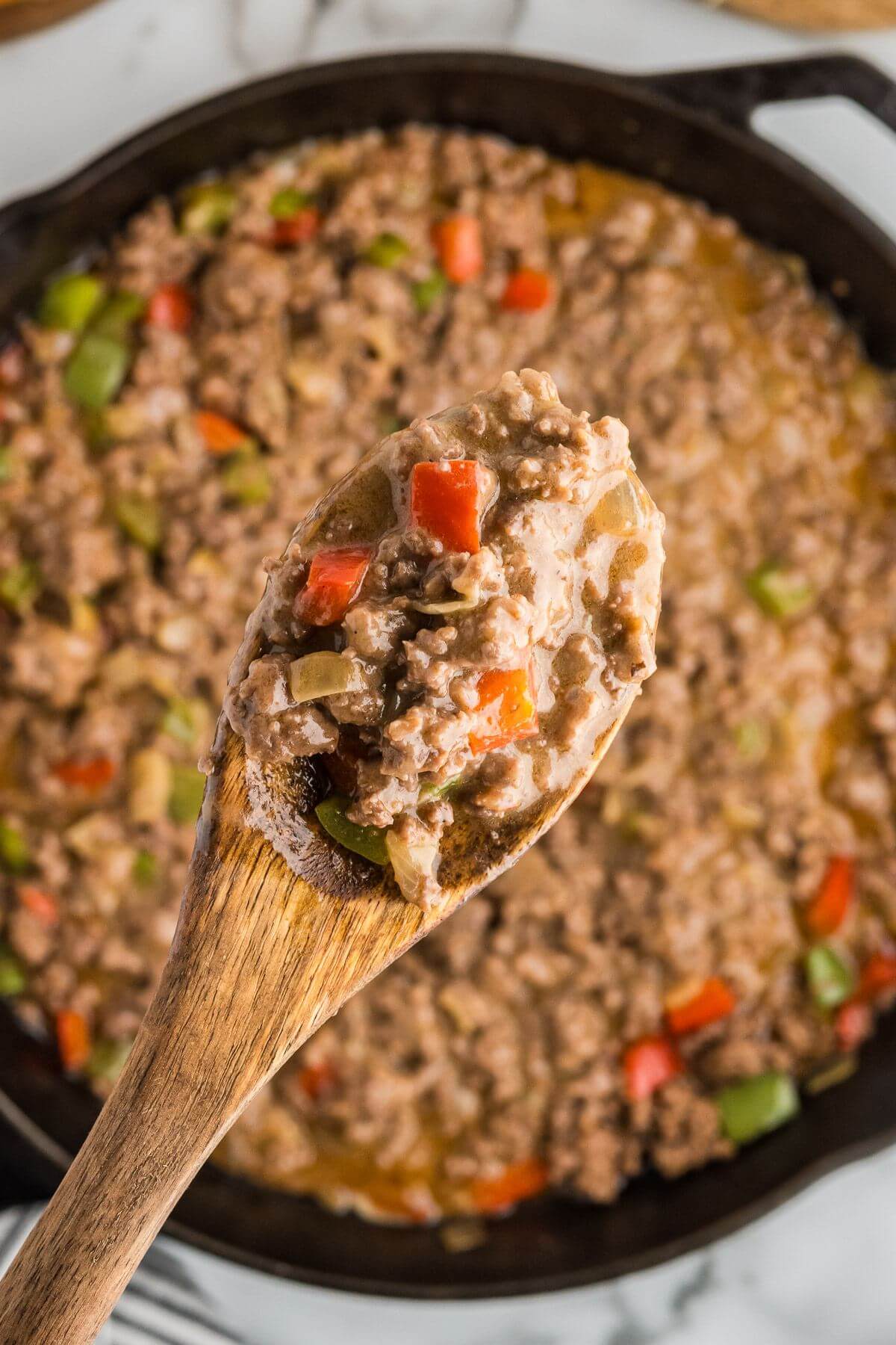 A wooden spoon holds up the saucy meat and peppers above the skillet.