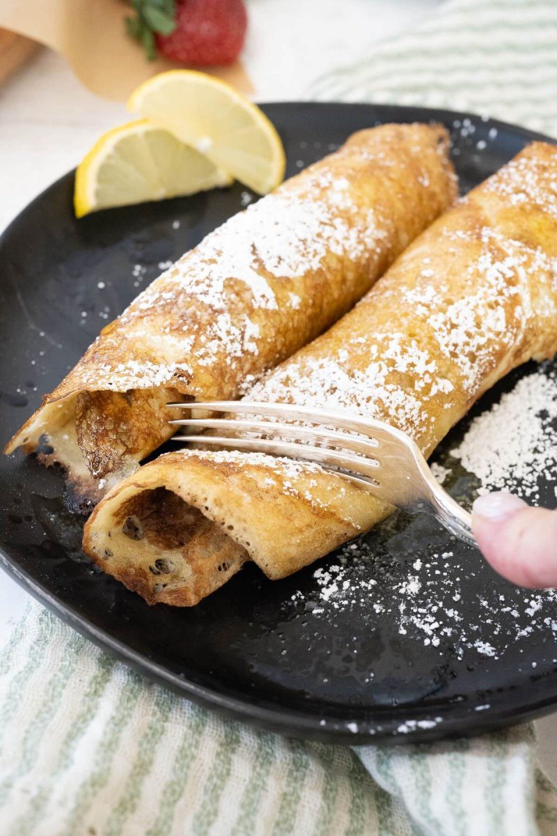 A hand uses a fork to cut into one of two sugared, rolled up pancakes.