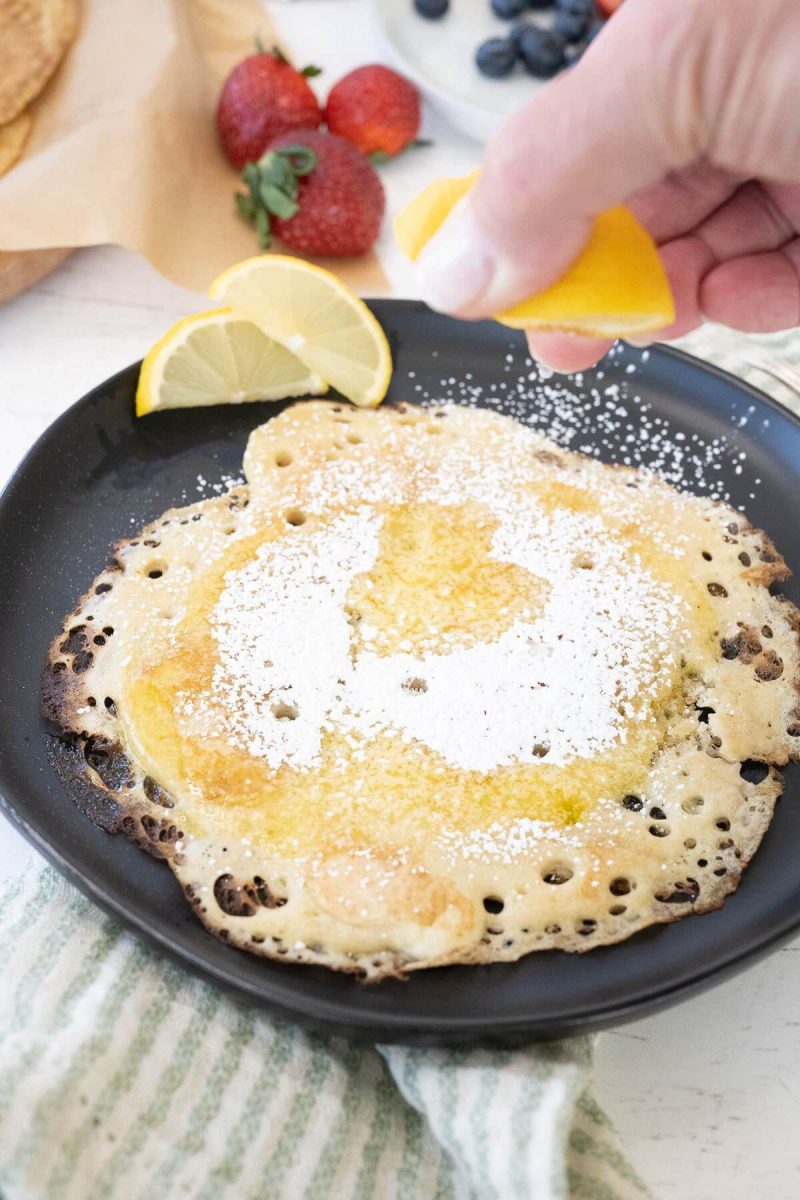 A hand squeezes a lemon slice over a buttered and sugared pancake on a black plate.