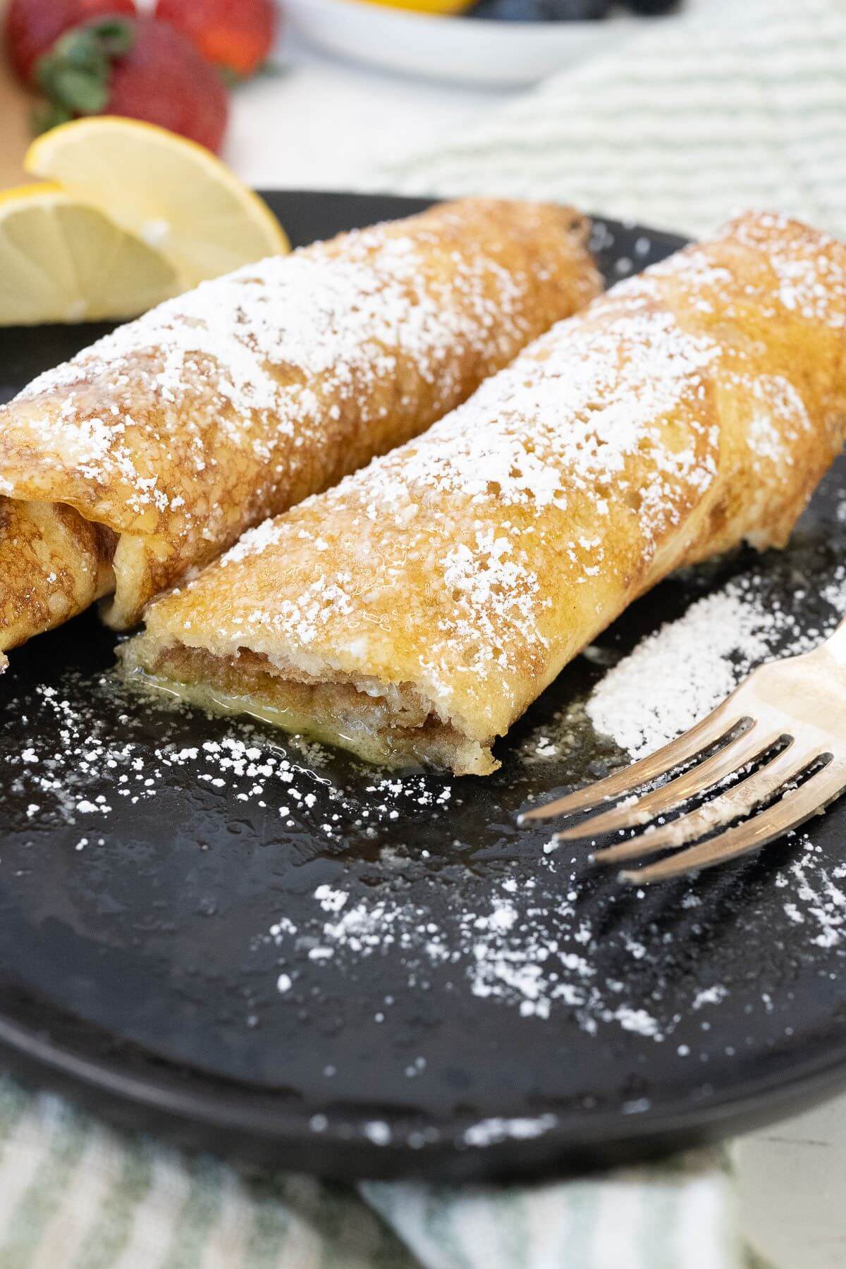 A fork lays next to two pancakes rolled up with powdered sugar on a plate.