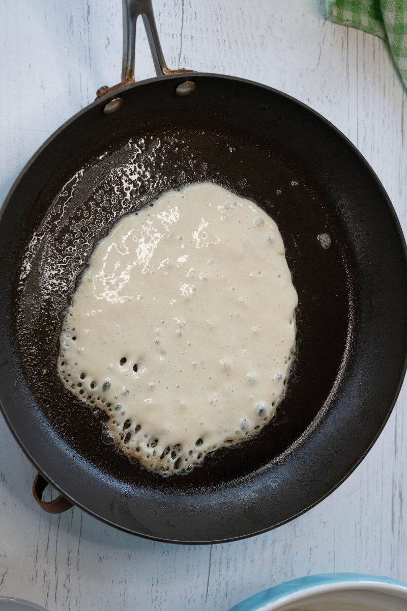 Pancake batter is starting to form bubbles while cooking in a cast iron pan.